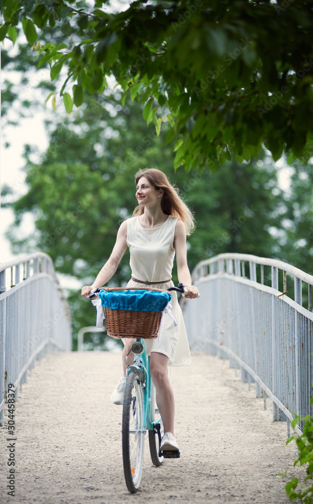 Carefree girl riding a blue bicycle with a basket in front, at the park having fun on summer day. Girl dressed in an airy light dress rides on the bridge in the background of lush greenery