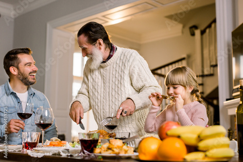 Portrait of happy old grandfather cutting turkey at festive table with his son and granddaughter eating meat