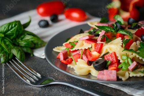 Traditional salad with pasta farfalle, ham, pepper and herbs on a dark plate on a dark background copy space