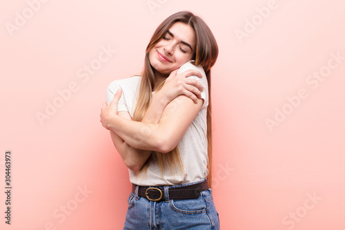 young pretty woman feeling in love, smiling, cuddling and hugging self, staying single, being selfish and egocentric against pink wall photo