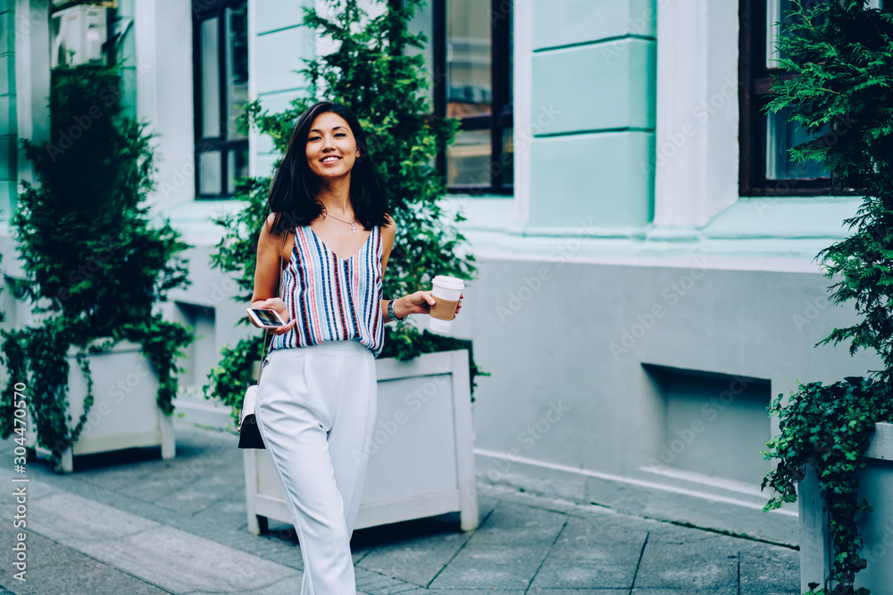 Half length portrait of cheerful female in trendy apparel walking on street with coffee to go,smiling asian girl looking at camera passing buildings in downtown enjoying free time walk on weekends