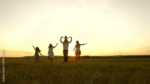 Happy young family with child run across field in sunset light. mother, father and little daughter with sisters walking in park. Children, dad and mom play in meadow in sunshine.