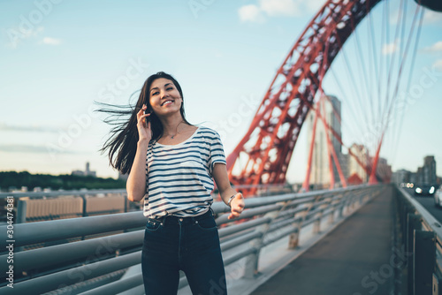 Half length portrait of cheerful hipster girl with modern cellphone device in hand posing at city urban setting and phoning to customer service for online consultancy, happy woman communicating