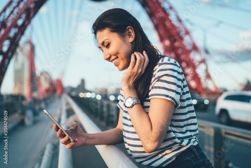 Side view of cheerful hipster blogger using mobile browsing for checking online incoming messages on social website, millennial Asian woman installing smartphone application for networking