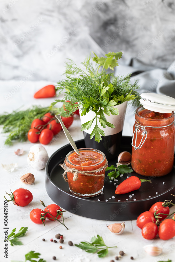A sauce of fresh red tomatoes in a glass jar on a black plate. Sprig of fresh cherry tomatoes, garlic, hot pepper, dill and parsley on a white table. A can of homemade ketchup.Close up A sauce of fresh red tomatoes in a glass jar on a black plate. Sprig of fresh cherry tomatoes, garlic, hot pepper, dill and parsley on a white table. A can of homemade ketchup.Close up