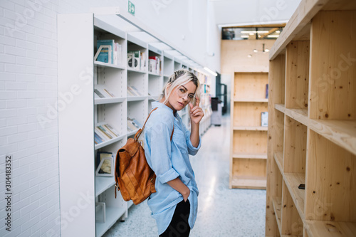 Half length portrait of confident caucasian female student dressed in stylish apparel looking at camera during spending leisure time in school library for education, young woman with backpack indoors