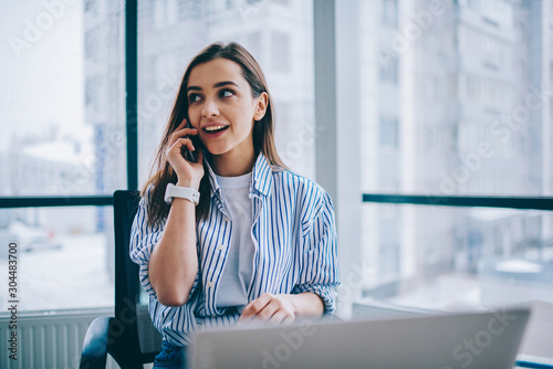 Cheerful young woman in casual wear having telephone conversation sitting at working place near laptop in office, smiling female freelancer calling to colleague for share good news about business.