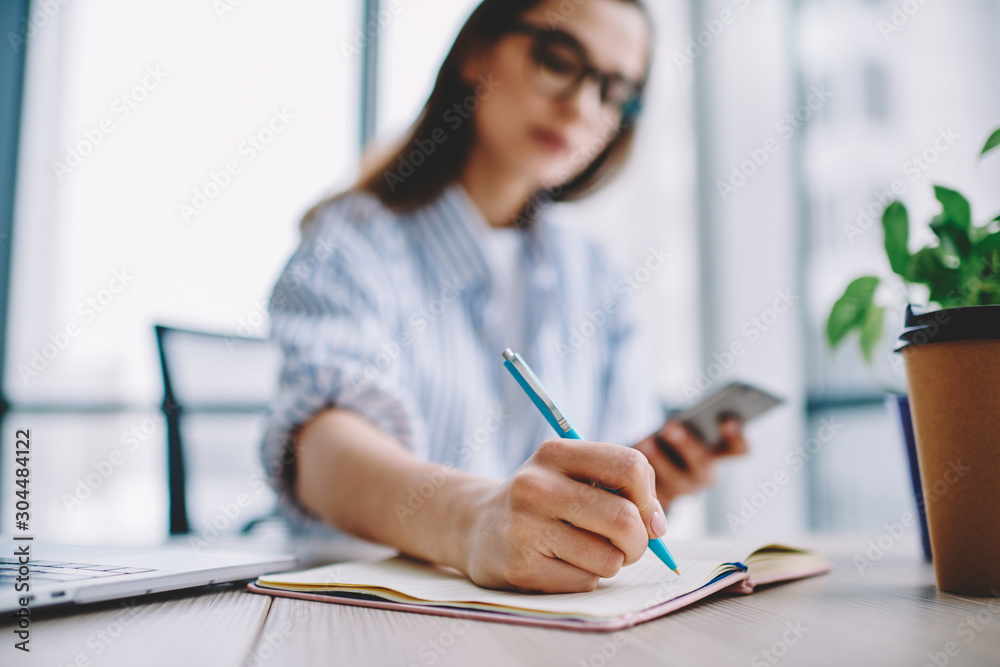 Selective focus on handwriting in notebook during creation of article for publication in blog, young woman on blurred background holding smartphone while making notes of creative ideas in office. Selective focus on handwriting in notebook during creation of article for publication in blog, young woman on blurred background holding smartphone while making notes of creative ideas in office.