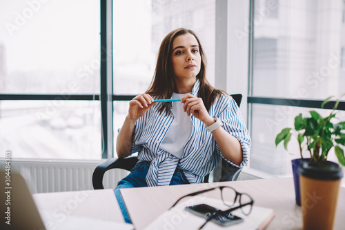 Serious brunette young woman sitting at desktop looking away pondering on idea for working project, creative female designer puzzled on business spending time at loft interior office holding pen. photo