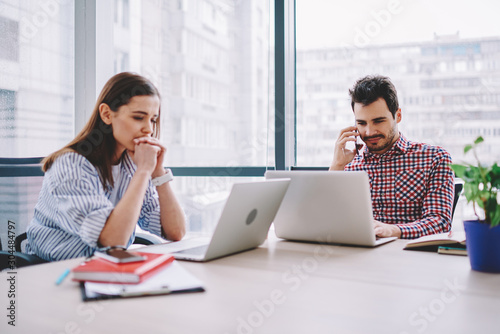 Concentrated male and female employees using laptop computers for making research in office,man talking on mobile phone during working process sitting at desktop with woman colleague read news online.