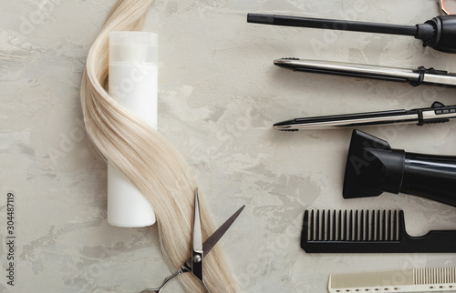 Flat lay composition with Hairdresser tools and white bottle of cosmetic product, blonde hair on gray background. Shampoo mockup. Space for design. Hairdresser service. Natural cosmetics concept.