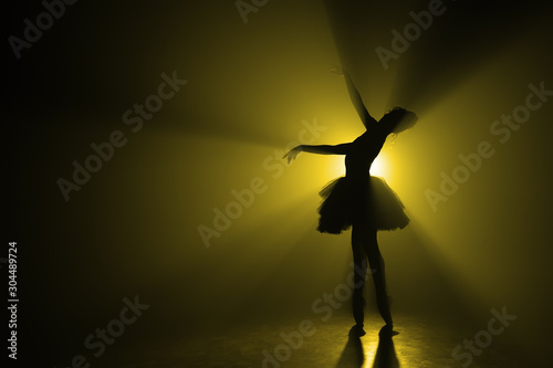 Copy space. Silhouette of dramatic girl dancing ballet in tutu on stage in front of spotlight with colored yellow neon light. Volumetric painting, smoke scene.