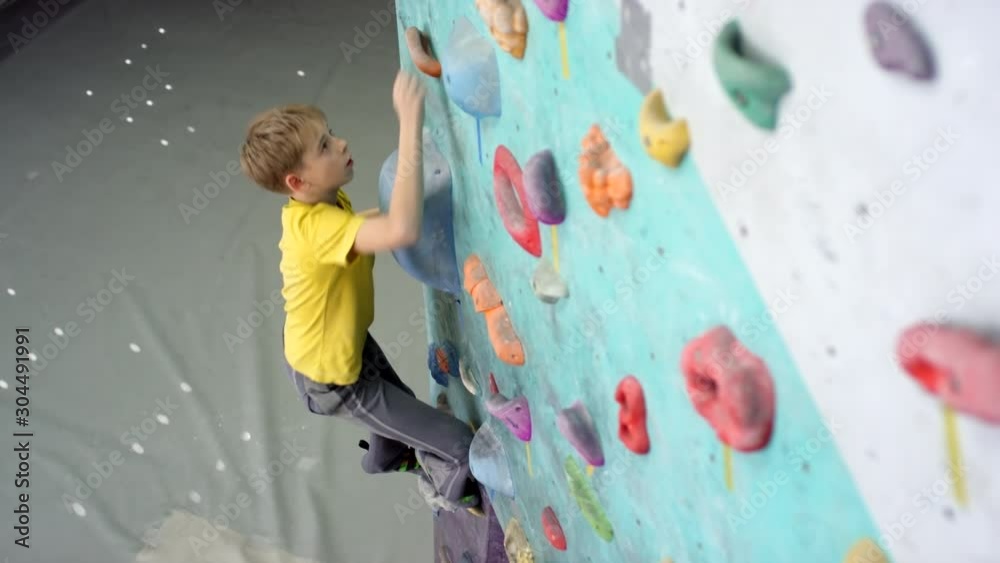 Tracking shot of 13-year-old blond Caucasian boy in yellow t-shirt, pants and shoes climbing up indoor vertical wall with manmade holds towards top, and padded crash mats on floor below Tracking shot of 13-year-old blond Caucasian boy in yellow t-shirt, pants and shoes climbing up indoor vertical wall with manmade holds towards top, and padded crash mats on floor below