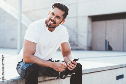 Positive sportsman dressed in trendy active wear looking away and smiling while resting on urban setting with modern cellphone in hands, happy male athlete feeling good after morning running