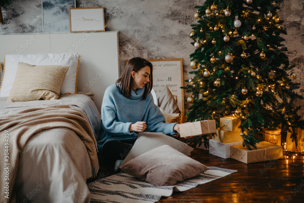 Fototapeta premium Young woman sits next to Christmas tree and holds box with gift.