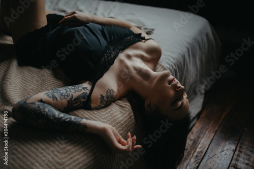 Young woman in black lingerie with tattoo on her arm lies on bed.
