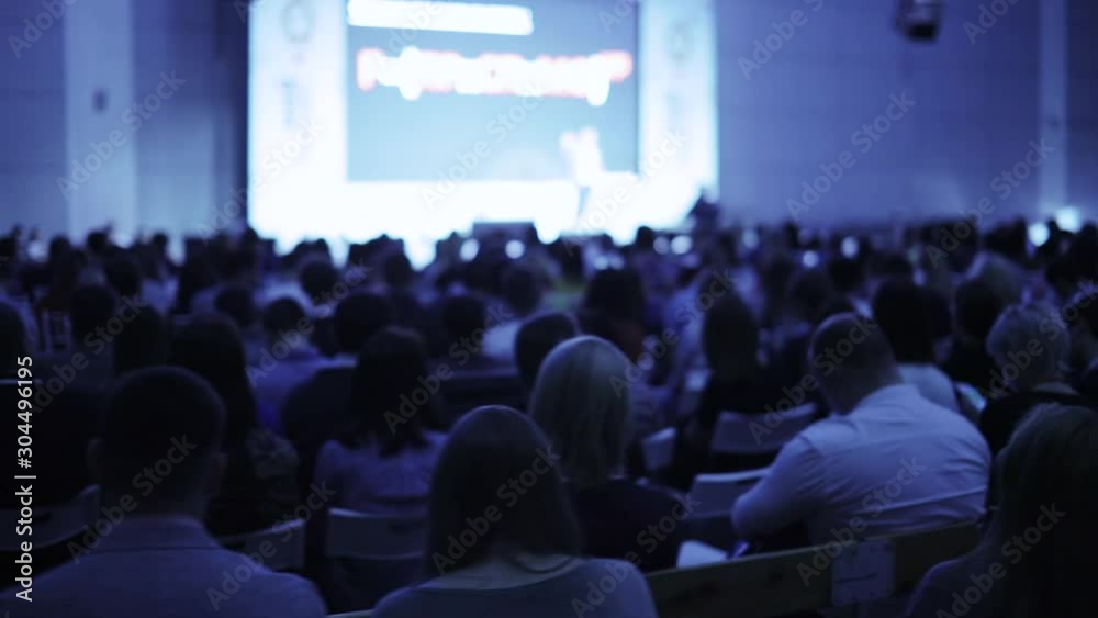 Unrecognizable businessman on stage talking microphone and showing presentation on screen to large audience sitting back to camera, blur background. Training, conference, public speech concept.