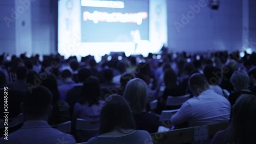 Unrecognizable businessman on stage talking microphone and showing presentation on screen to large audience sitting back to camera, blur background. Training, conference, public speech concept.