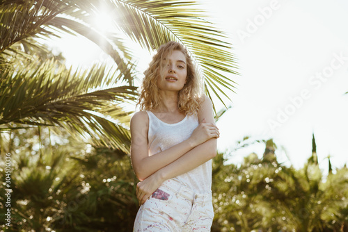 curly blonde clasped her arms around her shoulders and stands under the branches of a palm tree © nelen.ru