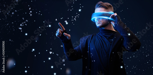 Beautiful young man using VR helmet while touching air in colorful lights. Guy in glasses of virtual reality with blue backlight over dark magic universe background. 