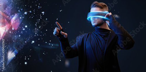 Beautiful young man using VR helmet while touching air in colorful lights. Guy in glasses of virtual reality with blue backlight over dark magic universe background. 