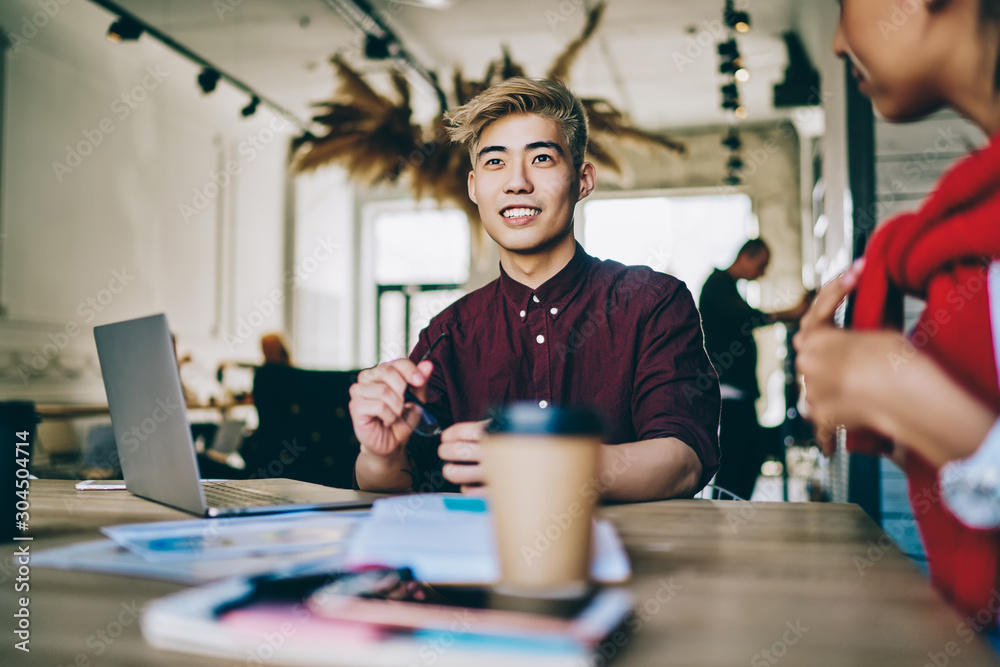 Cheerful smiling asian hipster student sitting at table with female colleague and feeling good from collaboration indoors, successful friends learning in coworking space using laptop computer Cheerful smiling asian hipster student sitting at table with female colleague and feeling good from collaboration indoors, successful friends learning in coworking space using laptop computer