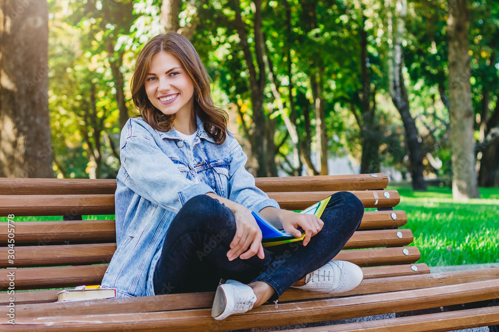 Obraz premium Young girl student sits on a bench in the park and holds books, notebooks. Girl teaches lessons in the park.