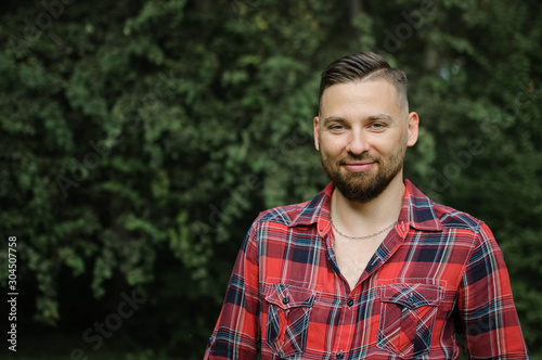Brutal bearded young man in red shirt is standing outdoors on green trees background. Hipster style concept.