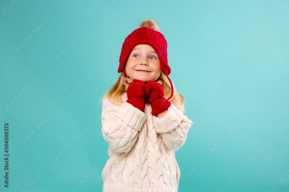 little blonde girl in a red knitted hat, mittens and a white sweater smiles on a blue background. winter clothes, space for text