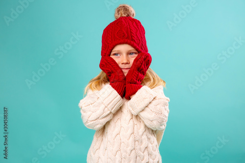 Konstfotografi little blonde girl in a red knitted hat, mittens and a white sweater smiles on a blue background