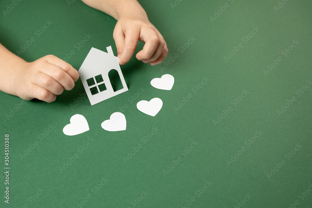 Child hands holding white paper house and heart on green background. Family home and real estate concept. Flat lay, copy space Child hands holding white paper house and heart on green background. Family home and real estate concept. Flat lay, copy space