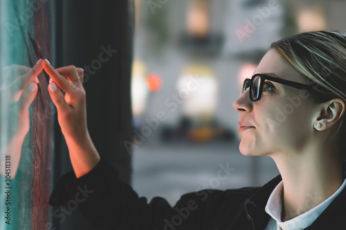 Smiling female standing at big display with advanced digital technology. Young woman touching with finger sensitive screen of interactive kiosk for find information while standing on street in evening