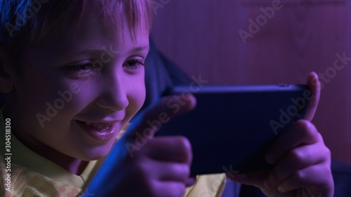 Boy plays games, in the evening. Gamer playing in the game using smartphone. Little caucasian kid holds mobile phone and plays online games. Joyful child spends time in the internet.