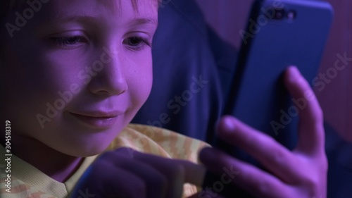 Child with smartphone, 8 years old. Boy typing text messages. Little boy looks to the screen cell phone in the evening. Caucasian kid holds phone and typing text. Child spends time in the internet