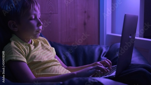 Child with computer at home, 8 years. Boy typing text message on keyboard. Little boy looks to the screen laptop. Caucasian kid holds notebook and typing text. Child spends time in the internet.