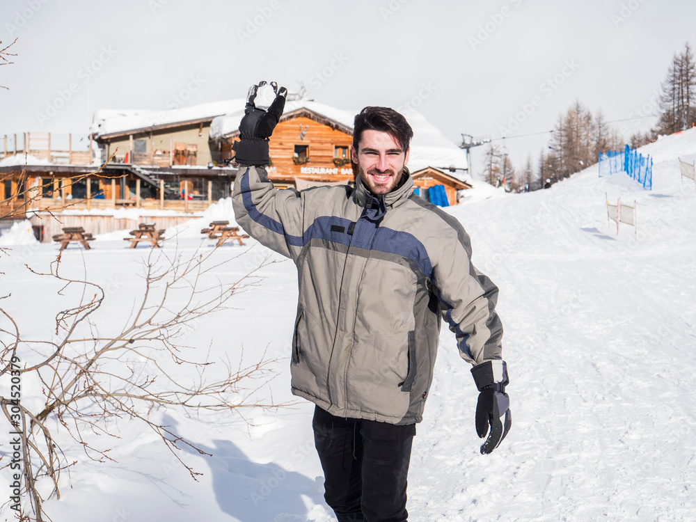 Cheerful handsome man in outerwear throwing snowball posing at camera in sunlight.