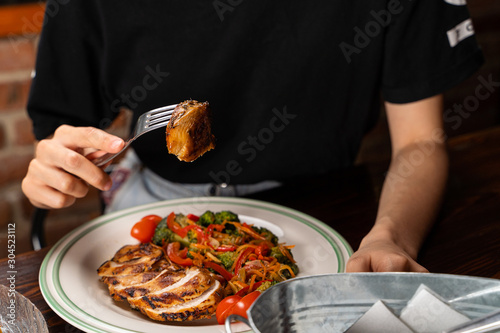 Young woman is eating grilled chicken breast with stewed vegetables:broccoli, carrot, tomatoes and bell pepper, alone with hands visible