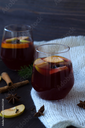 glasses of red mulled wine with orange, apple and cinnamon, anise star and warm white knitted plaid on a dark wooden background with green pine branch. winter still life. close up