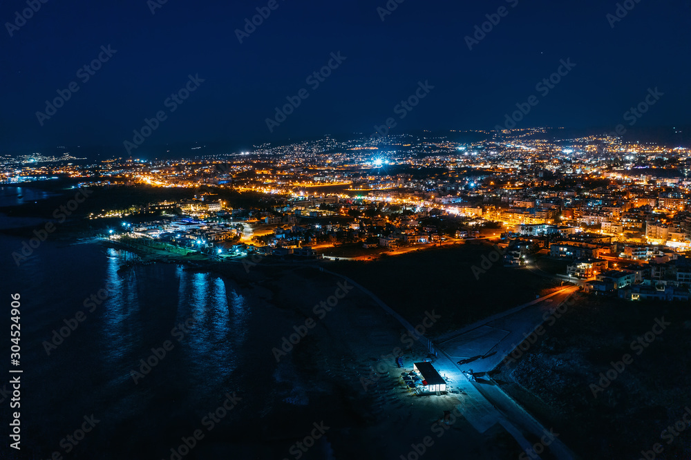 Aerial panoramic view of Paphos, Cyprus seaside from above at night from drone. Beautiful evening mediterranean seascape with illuminated buildings and reflections of lights in sea water.