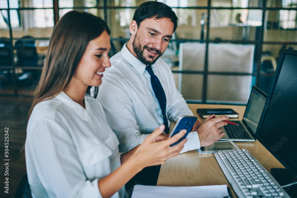 Obraz premium Positive male and female employees watching funny video online via smartphone gadget sitting at desktop with computers, successful colleagues enjoying communication togetherness reading news