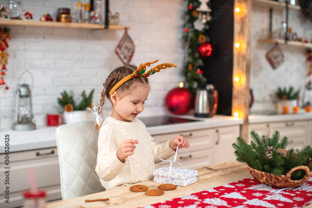 cute baby in light sweater and headband with reindeer horns sitting at kitchen table smiling opens gift box cute baby in light sweater and headband with reindeer horns sitting at kitchen table smiling opens gift box