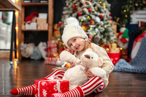 cute girl child 4 years in a light sweater and hat with pumponchik, sitting on the dark floor smiling wearing a plush toy hat Santa Claus.