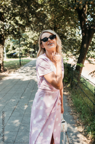 Beautiful stylish blonde woman in polka dotted midi dress in city summer park. She is happy and enjoying walk in sunny day. She is wearing small white bag and black sunglasses.  