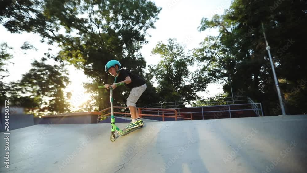 Concentrated boy stands on grey ramp edge and rides scooter down pool bank from orange handrails extreme slow motion	