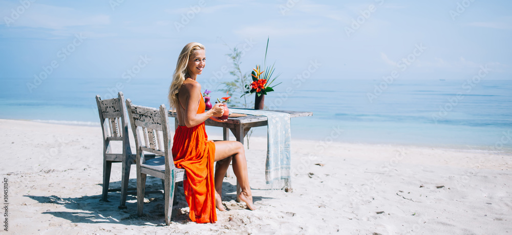 Adult woman drinking fresh beverage while sitting at table on sea sand beach