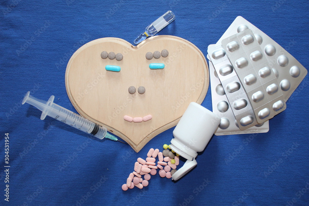 Big heart on a blue background. A smiley face is laid on it with pills. At  the top of the ampoule for injection, next to the scattered tablets and  syringe. Stock Photo