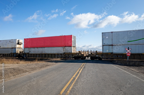 Eastern Alberta, Canada - Oct 23 2019: Freight train with cargo containers passing railroad crossing with stop road sign and asphalt road in the middle of prairie, Canadian National 