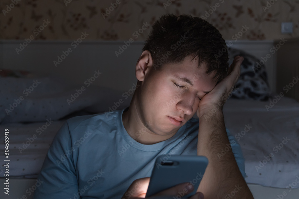Adult sleepy man awake late at night in bed surfing in web, can not fall asleep/ sleepy tired, social media addiction, dependency on cell phone, half-closed tired eyes, insomnia, sleeplessness concept Adult sleepy man awake late at night in bed surfing in web, can not fall asleep/ sleepy tired, social media addiction, dependency on cell phone, half-closed tired eyes, insomnia, sleeplessness concept