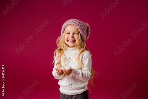 little blonde girl in a knitted hat and sweater catches the snow and laughs. winter clothes. isolate on red background, space for text
