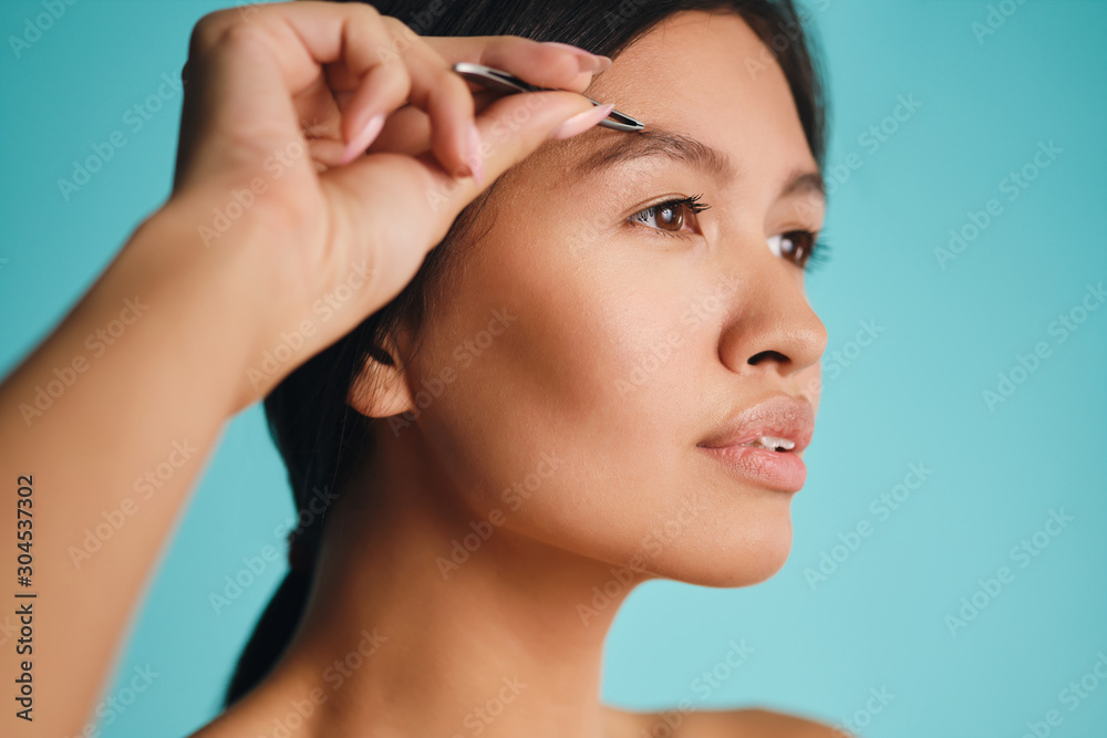 Side view of beautiful Asian brunette woman dreamily plucking eyebrows over colorful background isolated